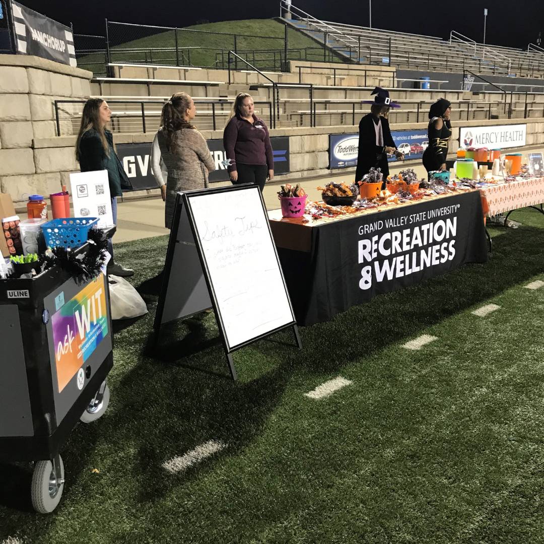 Image of students standing behind a Recreation & Wellness table at Halloween event in Lubbers stadium.